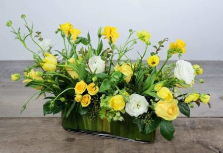 A cheerful centerpiece for your gathering. Buttercream roses, butterfly ranunculus, tulips, lisiantus, freesias with seasonal foliage designed in a glass rectangular vase.