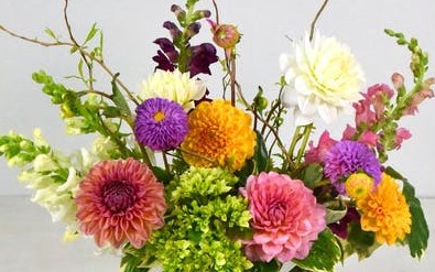 The whimsy of late summer is on display with colorful dahlias and snapdragons. Designed for a table or desk in a slender moon glass vase accented with curly willow and seasonal foliage.